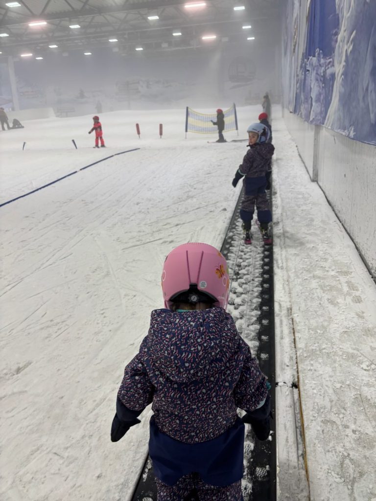 Kind fährt auf dem Zauberteppich einer Skihalle nach oben, von hinten fotografiert